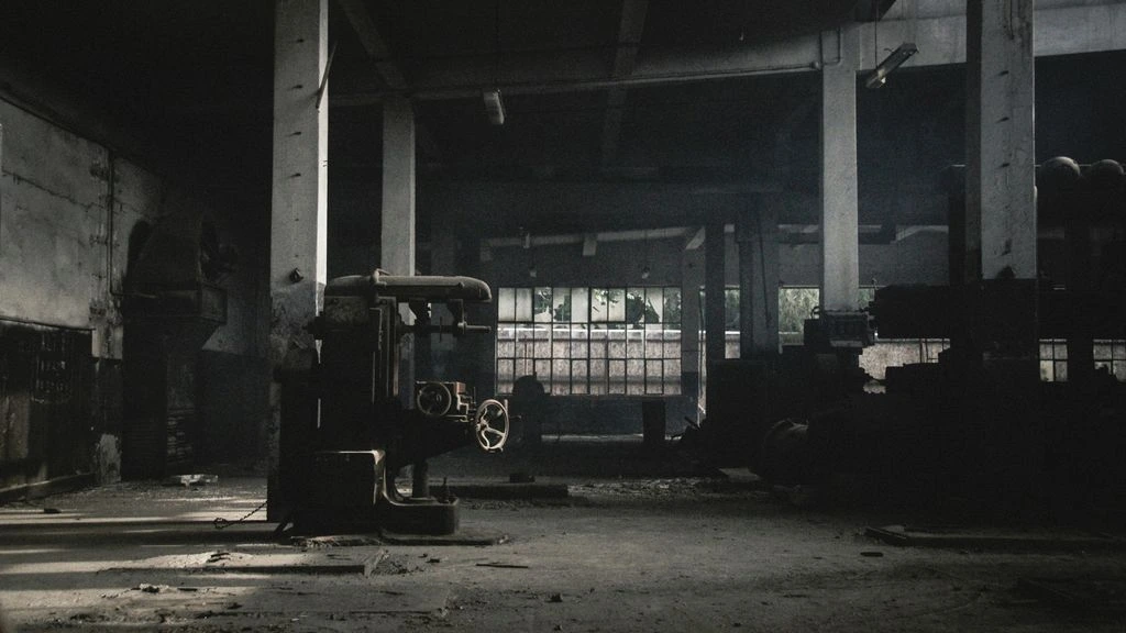 A moody industrial warehouse interior lit by a single warm tungsten key light, with a film crew working under soft backlight.